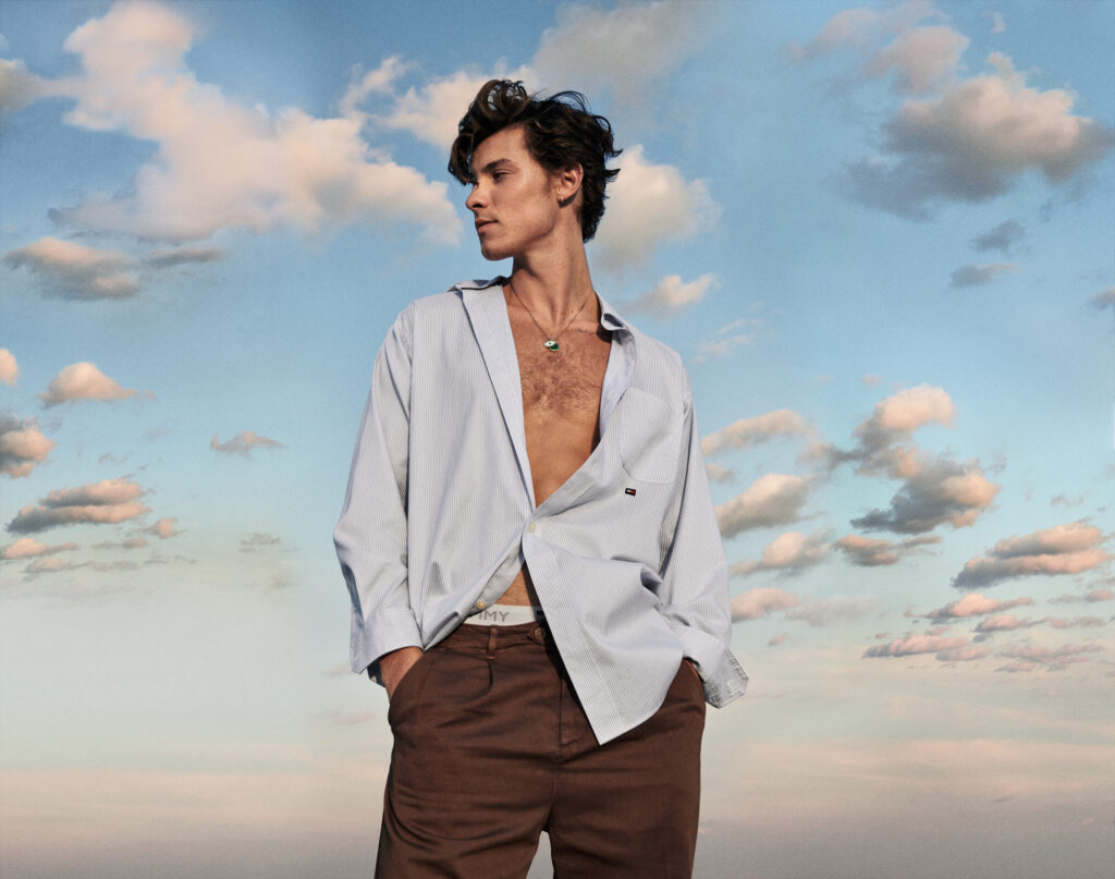 Shawn Mendes wears unbuttoned, baggy shirt, posing against a blue sky