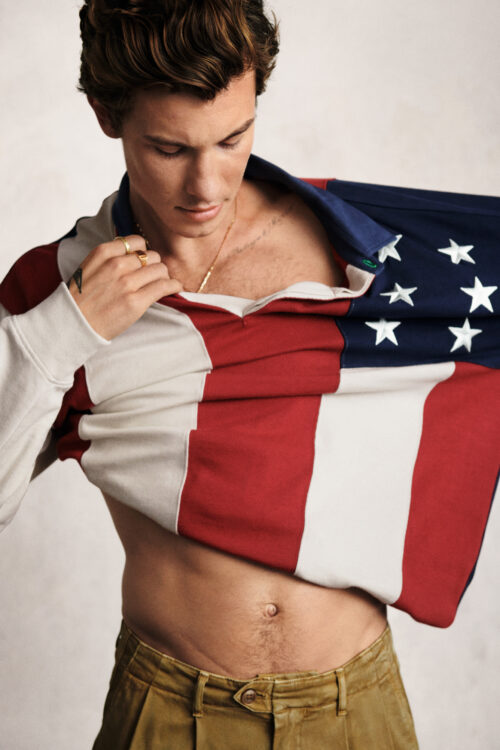 Shawn Mendes pulls at a stars and stripes rugby top to reveal some skin