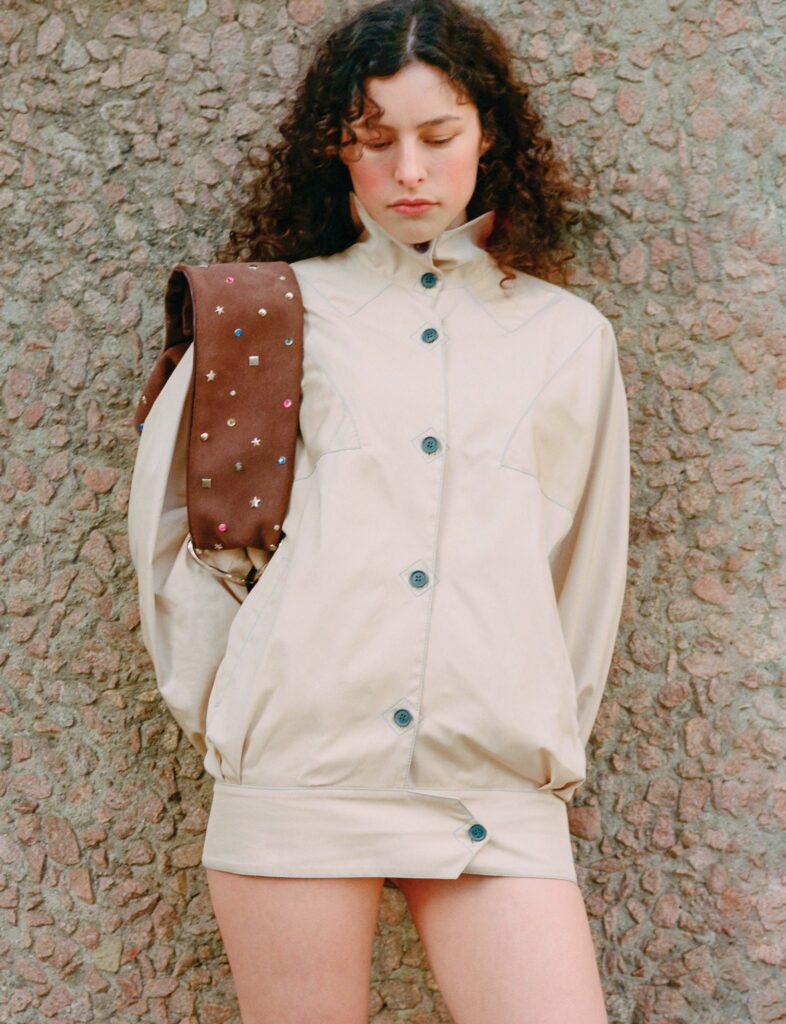 Torso shot of a model against a concrete wall wearing beige jacket with a thick-strapped, embellished brown bag