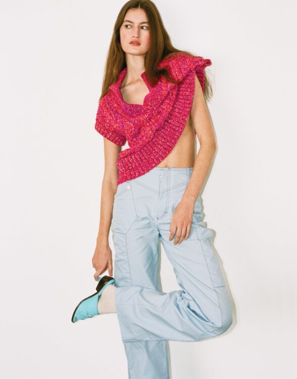 Full-length shot of model grabbing heel of shoe, wearing pink knitted sweater and washed-out blue cargos