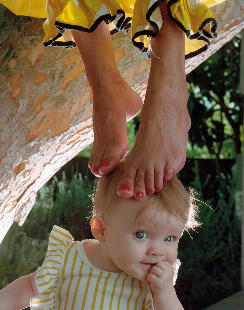 Feet hanging from the top of the photograph touch the head of a baby who looks towards the camera